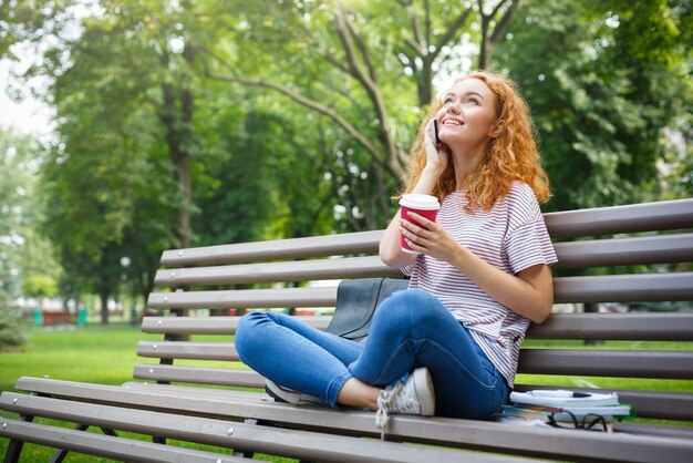 Joven estudiante pelirroja feliz hablando por teléfono inteligente, bebiendo café, sosteniendo la computadora portátil en la rodilla, sentado al aire libre en el banco en el parque con café