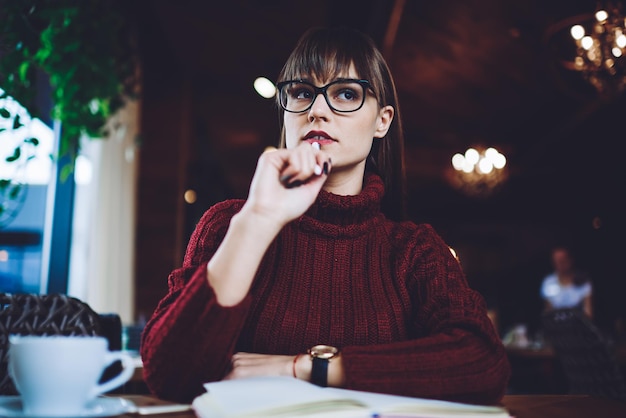 Una joven estudiante pensativa reflexionando sobre la idea de sentarse con un cuaderno de conocimientos en una cafetería