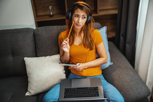 Una joven estudiante pensativa sentada en el sofá, usando su laptop y auriculares para ver lecciones en línea y aprender desde casa.