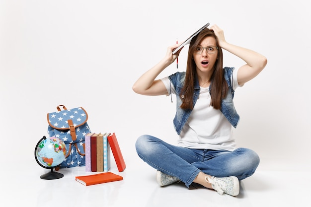 Foto joven estudiante preocupada y desconcertada con gafas sostiene un lápiz, un cuaderno aferrado a la cabeza se sienta cerca de la mochila del globo, libros escolares aislados en la pared blanca