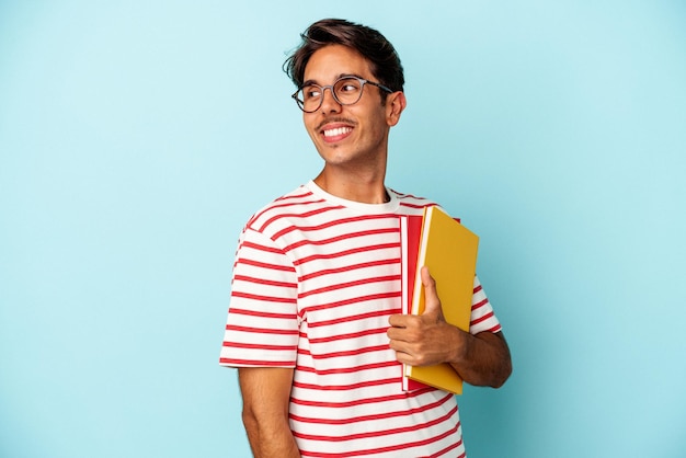 Un joven estudiante de raza mixta que sostiene libros aislados de fondo azul parece sonriente, alegre y agradable.