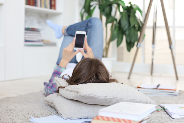 Joven estudiante revisando las redes sociales antes de volver a estudiar, acostada en el piso contra el acogedor interior doméstico, rodeada de una pila de libros.