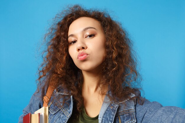 Joven estudiante en ropa de mezclilla y mochila sostiene libros, envía un beso y hace una foto selfie aislada en la pared azul