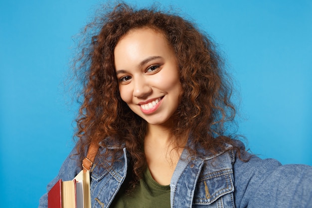 Joven estudiante en ropa de mezclilla y mochila sostiene libros y hace selfie foto aislada en la pared azul