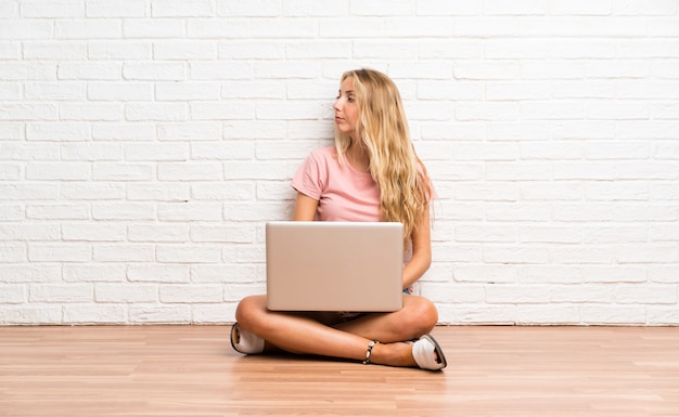 Joven estudiante rubia con una computadora portátil en el piso mirando hacia el lado