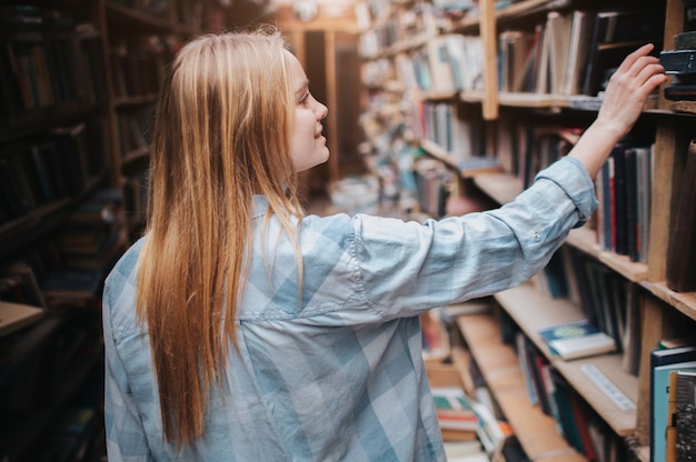 Joven estudiante rubia está eligiendo un libro de la estantería. Ella necesita encontrar el libro adecuado para prepararse para el examen. De cerca.