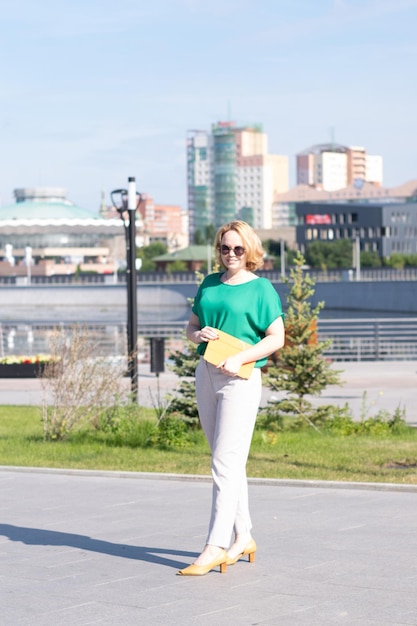 Una joven estudiante rubia feliz con gafas de sol caminando por una calle de la ciudad lleva una tableta digital naranja