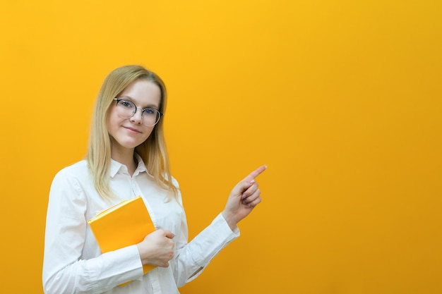 Joven estudiante rubia hermosa con gafas sostiene libros sobre un fondo amarillo