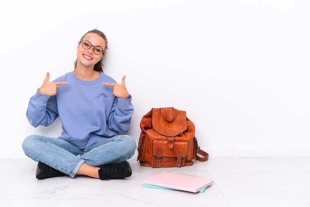 Joven estudiante sentada en el suelo aislada de fondo blanco orgullosa y satisfecha