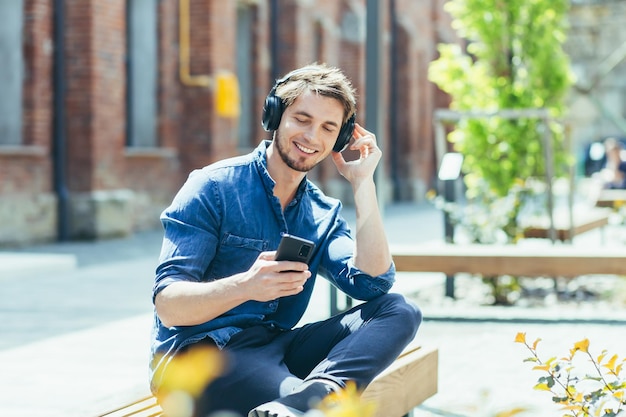 Joven estudiante sentado en el campus en un banco en la posición de loto escuchando una conferencia de audio de un audiolibro con auriculares sosteniendo un teléfono en sus manos Él sonríe