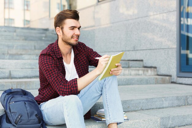 Joven estudiante sentado en las escaleras al aire libre, tomando notas, preparándose para los exámenes en la universidad o la universidad. educación, concepto de inspiración, espacio de copia.