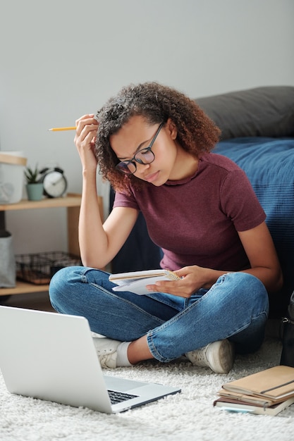 Joven estudiante seria con las piernas cruzadas sentada en el suelo junto a la cama frente a la computadora portátil y resding sus notas en el cuaderno en el entorno hogareño