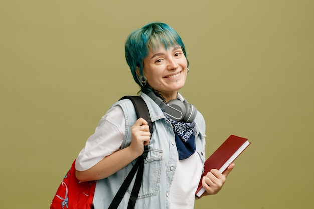 Joven estudiante sonriente con auriculares y pañuelo en el cuello y mochila de pie en la vista de perfil sosteniendo un cuaderno agarrando una mochila mirando la cámara aislada en un fondo verde oliva