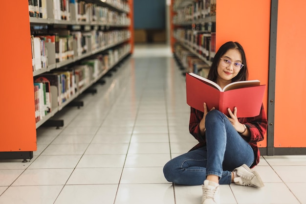 Joven estudiante sonriente con estilo informal sentado en el suelo junto a una estantería en la biblioteca