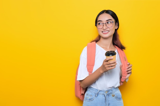 Una joven estudiante sonriente con mochila y gafas sosteniendo una taza de café para llevar aislada de fondo amarillo