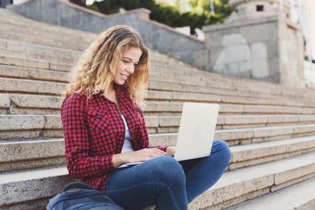 Joven estudiante sonriente sentada en las escaleras usando una laptop al aire libre durante el descanso descansando en el campus universitario. Concepto de tecnología, educación y trabajo remoto, espacio de copia