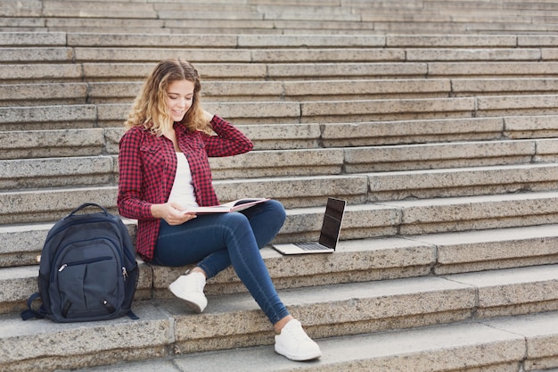 Joven estudiante sonriente sentada en las escaleras usando una laptop al aire libre durante el descanso, descansando en el campus universitario. Concepto de tecnología, educación y trabajo remoto, espacio de copia