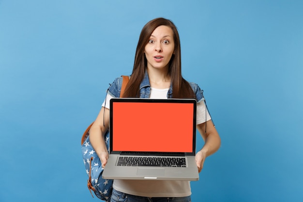 Joven estudiante sorprendida en ropa de mezclilla con mochila con ordenador portátil con pantalla en blanco vacía negra aislada sobre fondo azul. Educación en la universidad. Copie el espacio para publicidad.