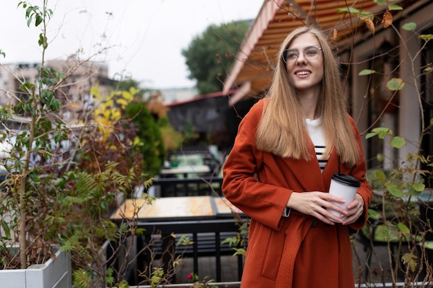 Una joven estudiante con una taza de café en las manos junto a un café callejero con una sonrisa en la cara