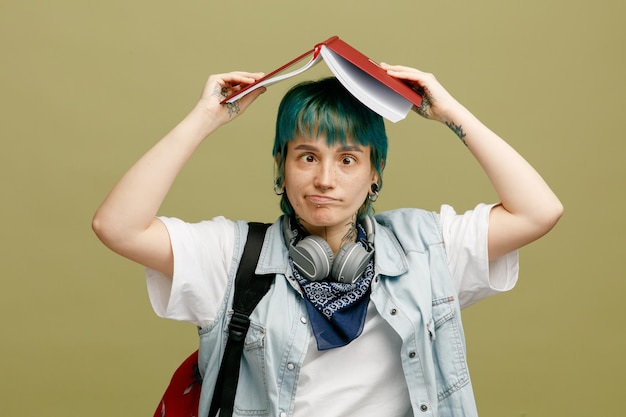 Una joven estudiante tonta con auriculares y pañuelo en el cuello y una mochila sosteniendo un cuaderno abierto sobre la cabeza mirando a la cámara cruzando los ojos aislada en un fondo verde oliva