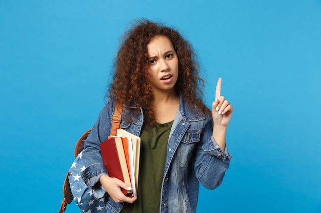 Joven estudiante triste en ropa de mezclilla y mochila tiene libros aislados en la pared azul