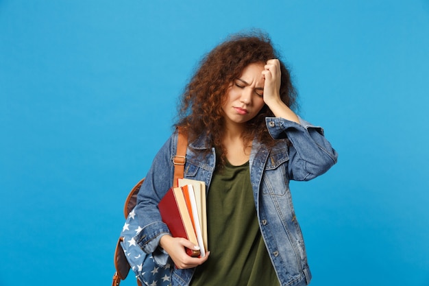 Foto joven estudiante triste en ropa de mezclilla y mochila tiene libros aislados en la pared azul