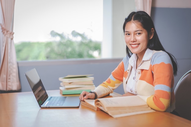 Joven estudiante universitaria asiática en uniforme de estudiante estudiando, leyendo un libro, computadora portátil en la biblioteca de la universidad o la universidad. Joven estudiante y educación de tutoría con un concepto de aprendizaje tecnológico