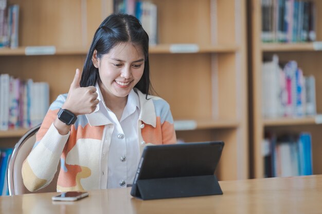 Joven estudiante universitaria asiática con uniforme de estudiante estudiando en línea, leyendo un libro, usando tableta digital o computadora portátil en la biblioteca de la universidad mientras el aula está restringida durante la pandemia de COVID-19