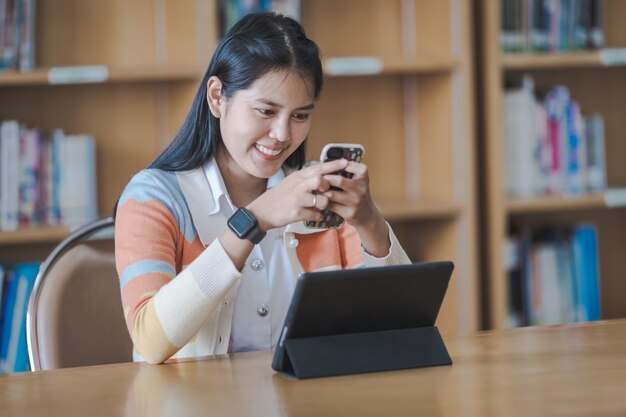 Joven estudiante universitaria asiática con uniforme de estudiante estudiando en línea, leyendo un libro, usando tableta digital o computadora portátil en la biblioteca de la universidad mientras el aula está restringida durante la pandemia de COVID-19