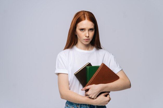 Una joven estudiante universitaria disgustada y triste sosteniendo un libro y mirando la cámara con un fondo gris aislado. Bonita modelo de dama pelirroja con ropa informal mostrando emocionalmente expresiones faciales.