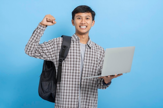 Joven estudiante universitario asiático con camisa de franela que lleva libros y mochila aislado sobre fondo azul.