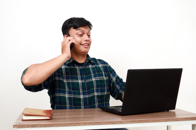 Joven estudiante universitario asiático sentado hablando por teléfono y usando una computadora portátil en el escritorio