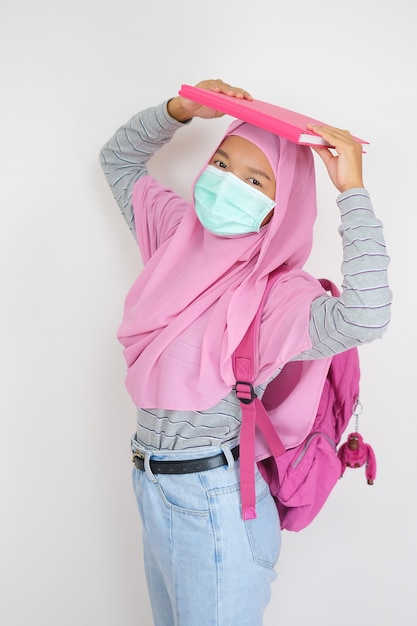 Joven estudiante usa hijab rosa y máscara sostiene libros sobre fondo blanco.
