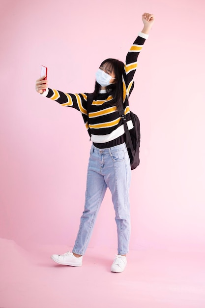La joven estudiante usa máscara y usa un teléfono inteligente con fondo rosa.
