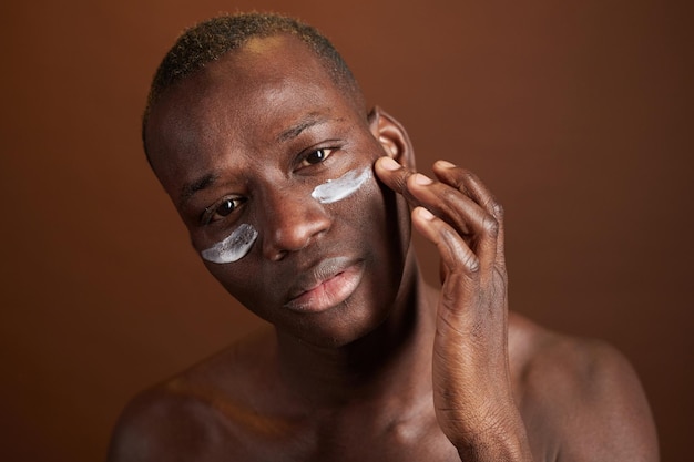 Joven de etnia africana mirándote mientras aplicas crema en el área debajo de los ojos