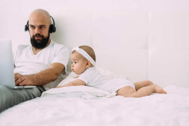 Un joven de etnia caucásica con auriculares trabaja en una laptop sentada en casa en la cama junto a su pequeña hija yace y mira