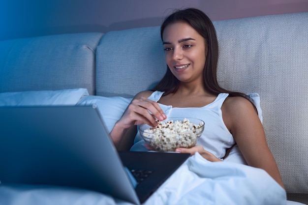 Una joven étnica sonriente acostada en la cama con un tazón de palomitas de maíz y viendo una laptop