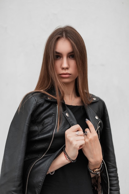 Joven europea con un elegante vestido endereza una elegante chaqueta de cuero negro cerca de un edificio blanco vintage. Modelo de moda chica muy hermosa sexy posando sobre un fondo blanco. Estilo de otoño.