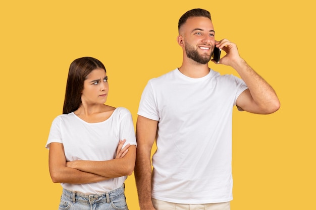 Una joven europea enojada con una camiseta blanca mira a un hombre feliz llamando por teléfono.