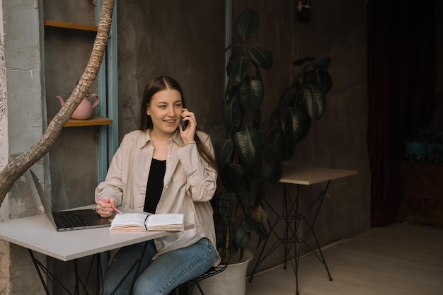Una joven europea independiente con cabello largo y oscuro en una camisa y jeans trabaja de forma remota en un café