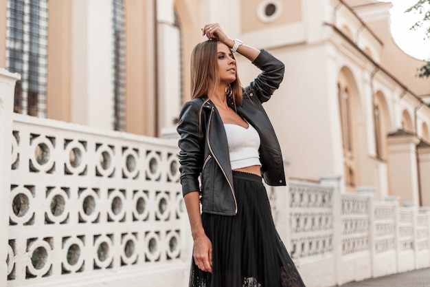 La joven europea de moda con una chaqueta de cuero negra de otoño a la moda con una falda larga elegante negra disfruta de un paseo por la ciudad. Modelo de chica bastante urbana posando cerca de una valla vintage en la calle