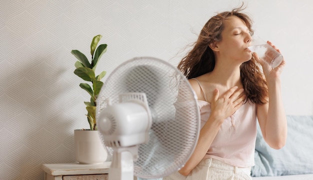 Foto joven europea se refresca en casa con la ayuda de un ventilador de aire acondicionado y bebe agua fría