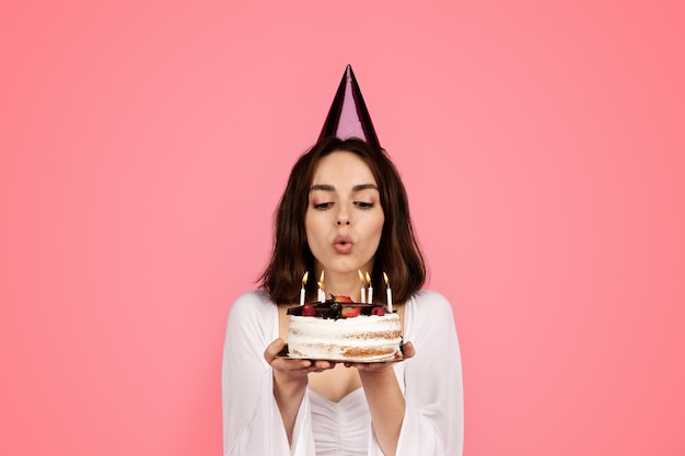 Una joven europea sonriente con sombrero sostiene un pastel que sopla velas y pide un deseo aislado de fondo rosa
