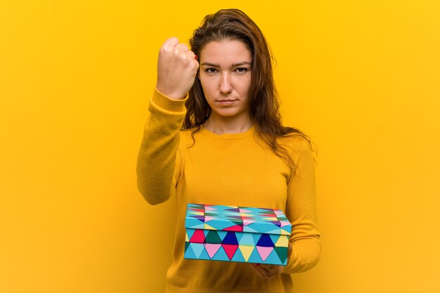 Joven europea sosteniendo un regalo mostrando el puño a la cámara, expresión facial agresiva.