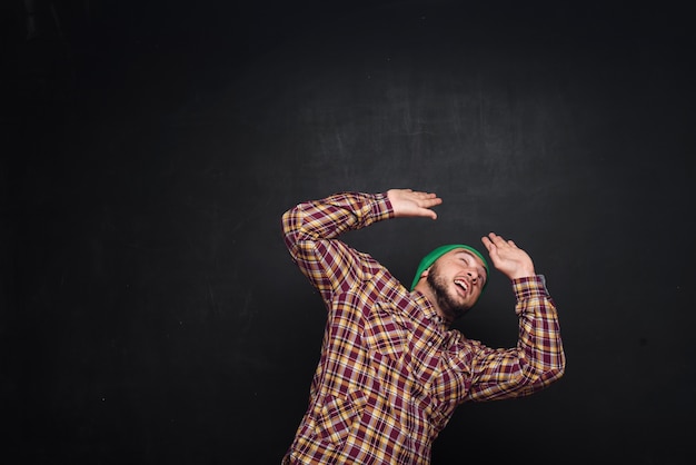 Joven europeo con barba en gorro de punto verde, parece sorprendido y desconcertado. Mostrando los dedos hacia arriba y el lado derecho. Fondo negro, espacio de copia en blanco para texto o publicidad