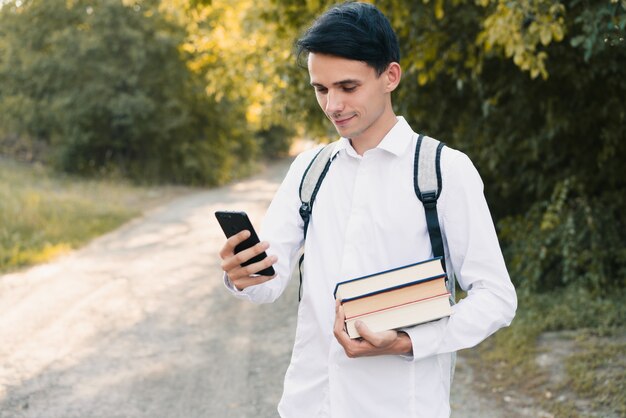 Un joven europeo con una camisa clásica blanca y una mochila gris en la espalda sostiene una pila de libros en la mano y mira a su teléfono, al aire libre. concepto de educación