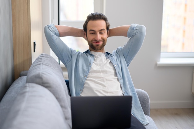 Joven europeo guapo con barba sentado en el sofá con computadora portátil en casa buscando en Internet viendo películas o series reunión remota el fin de semana relajado