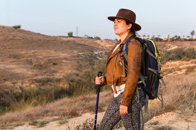 Una joven excursionista con una chaqueta de cuero, una mochila y un bastón de seguimiento camina por un campo