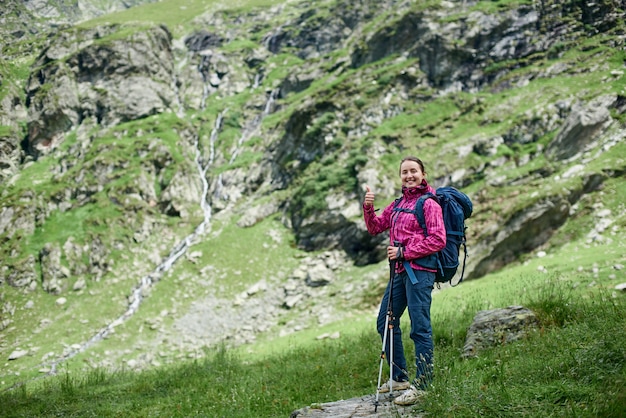 Joven excursionista feliz famele con bastones de trekking y una mochila está de pie sobre una piedra al pie de las montañas. Niña sonriendo a la cámara y mostrando los pulgares para arriba