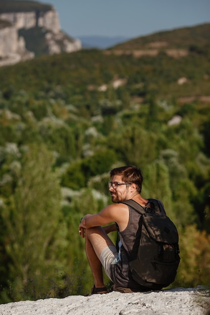 Joven excursionista con mochila relajándose en la cima de una montaña durante la tranquila puesta de sol de verano